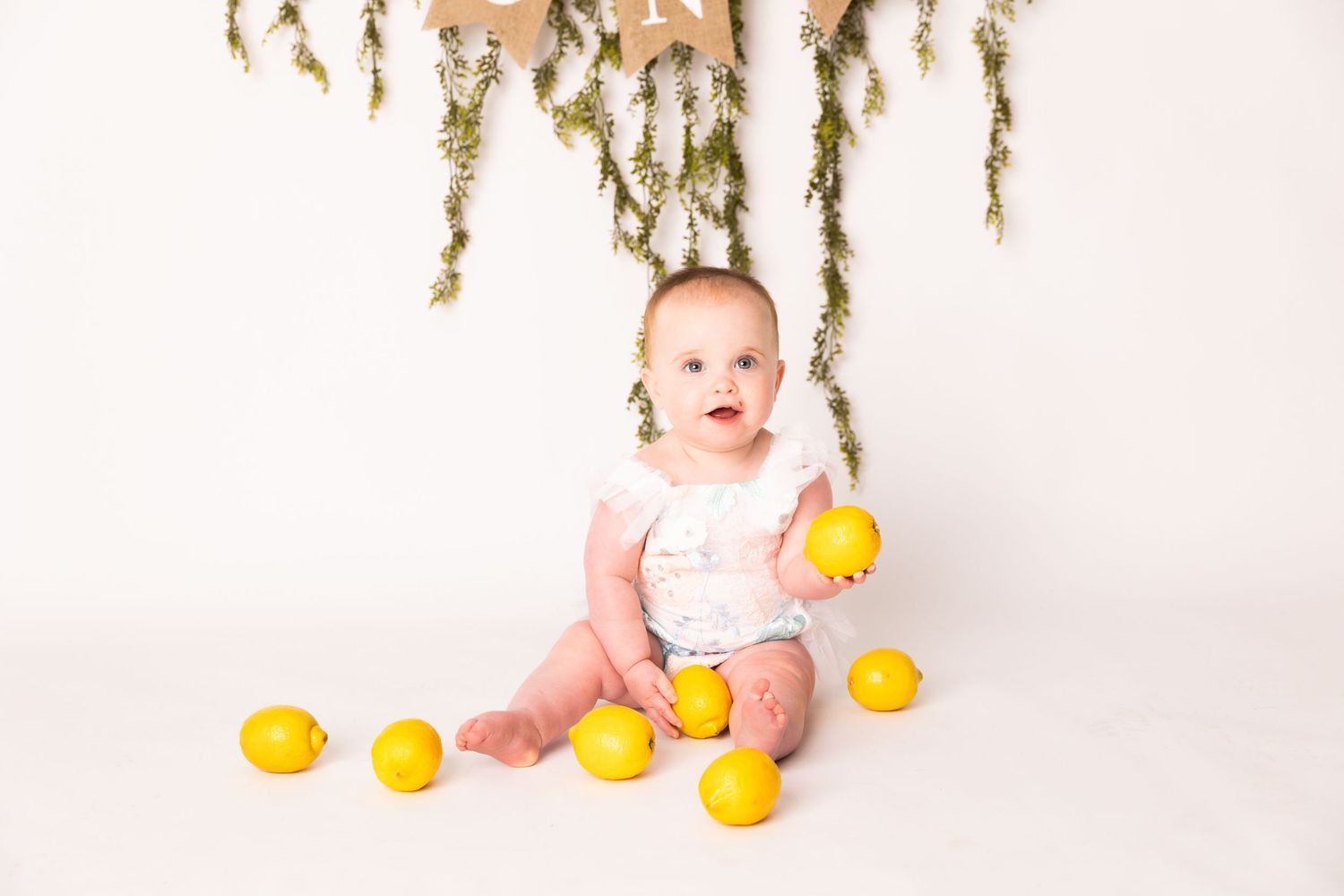 Baby in pink outfit sits surrounded by bright yellow lemons against white background with hanging dried flowers.