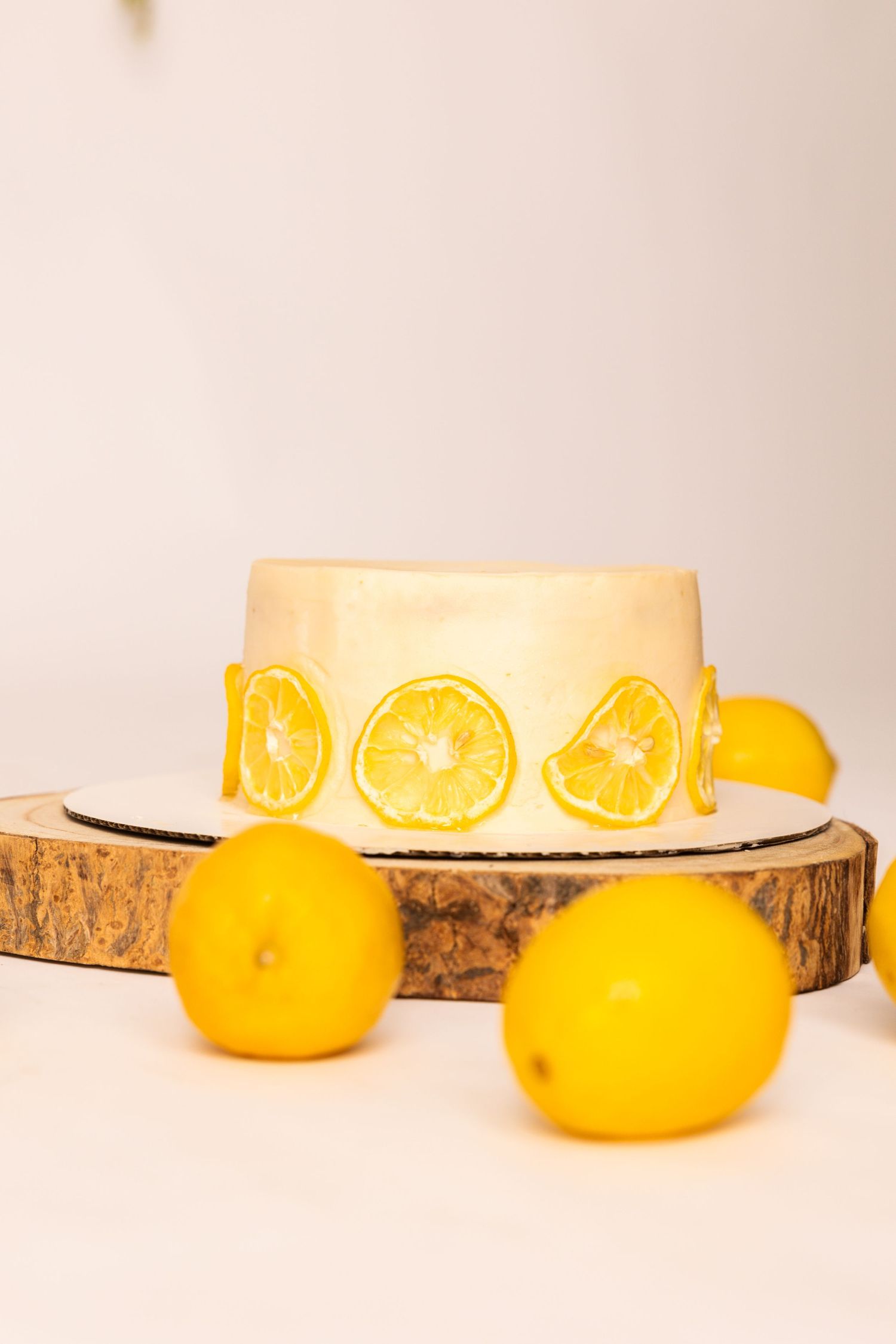 A simple white cake decorated with fresh lemon slices sits on a rustic wood slice with whole lemons alongside it.