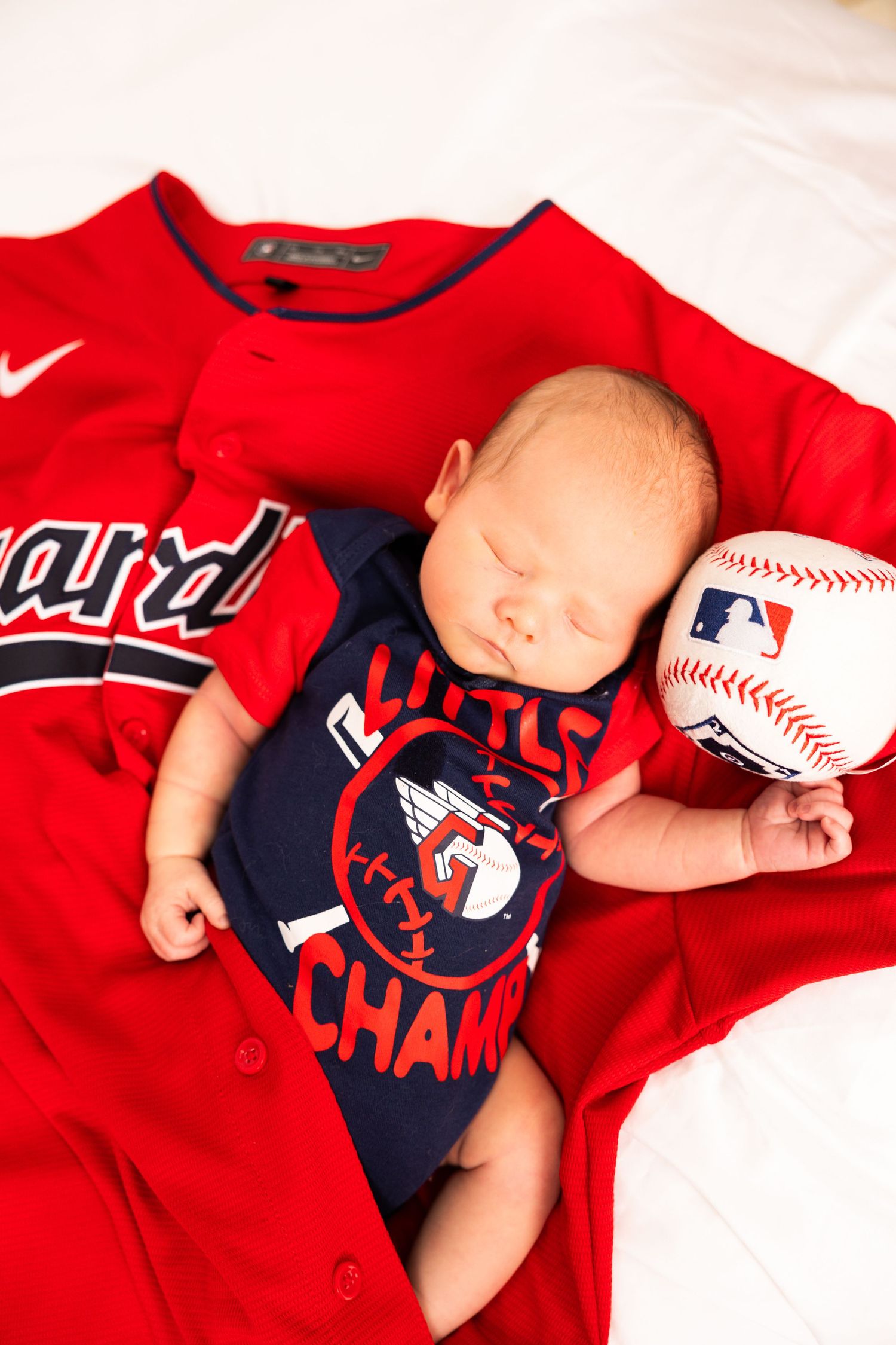 Sleeping newborn baby in Cleveland Guardians onesie nestled on red MLB jersey with baseball.
