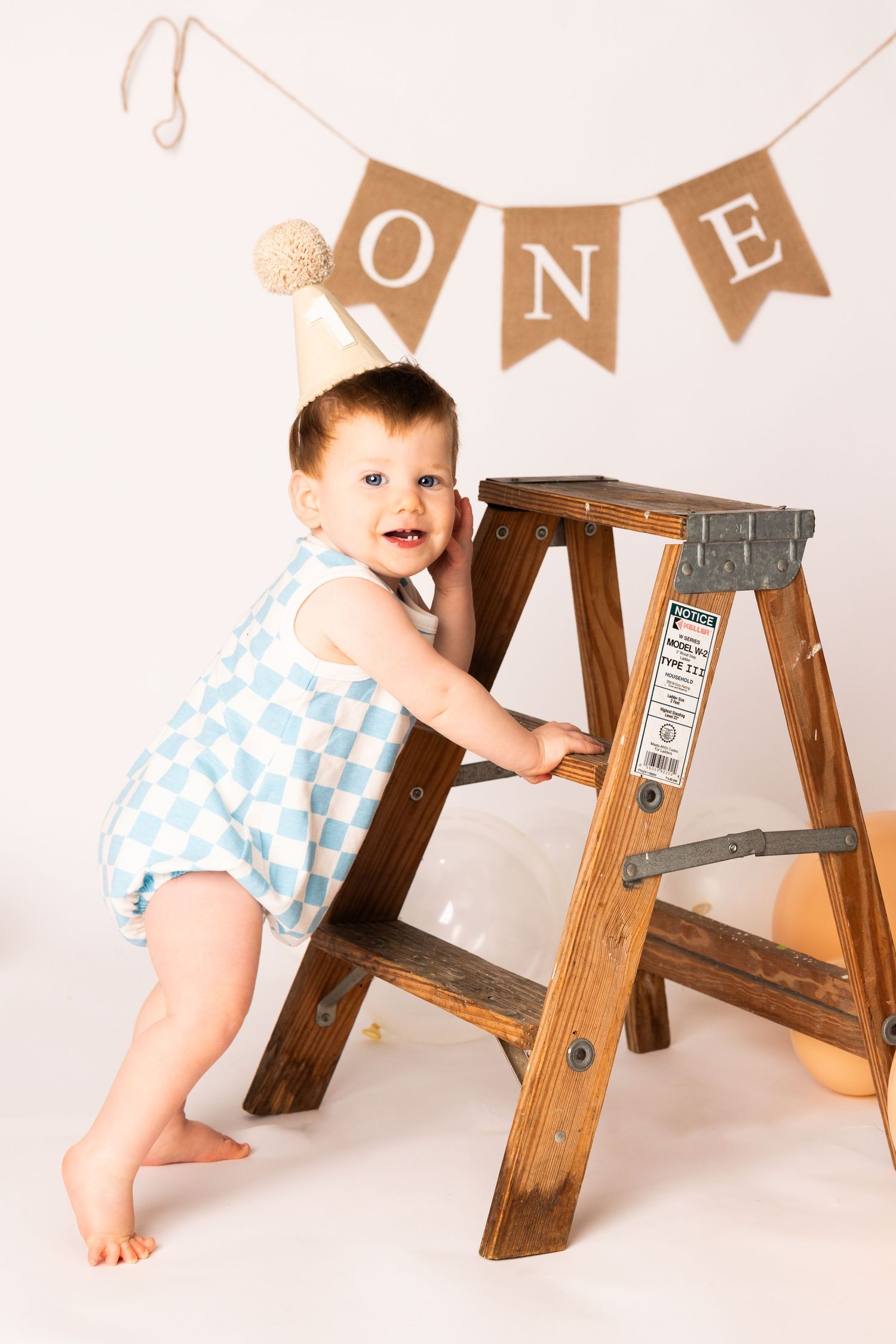 A one-year-old baby in a blue and white polka dot outfit plays with a wooden ladder during a milestone photo session.