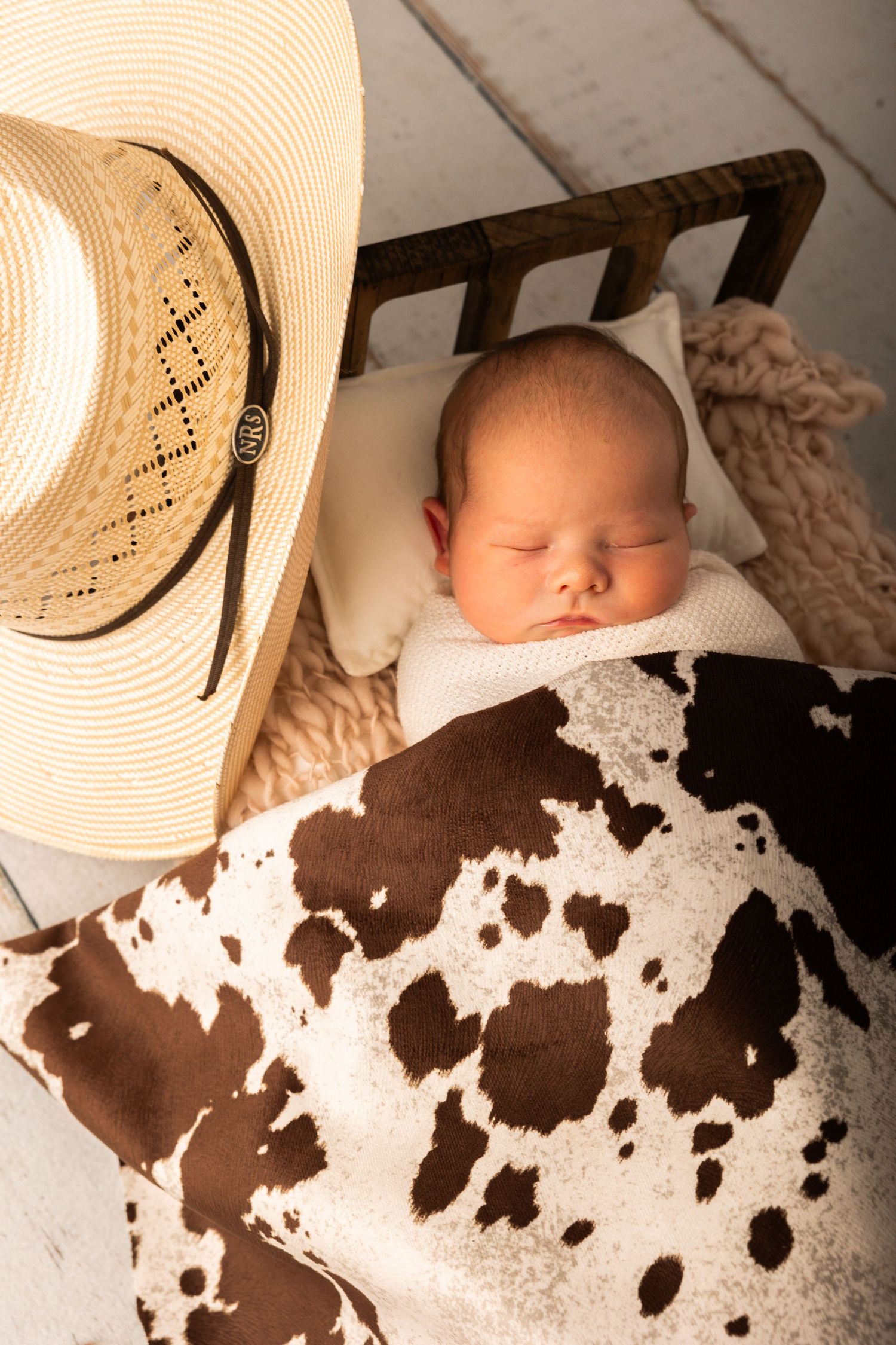 Peaceful newborn baby sleeping in rustic wooden bed with cowboy hat and cowhide print blanket.