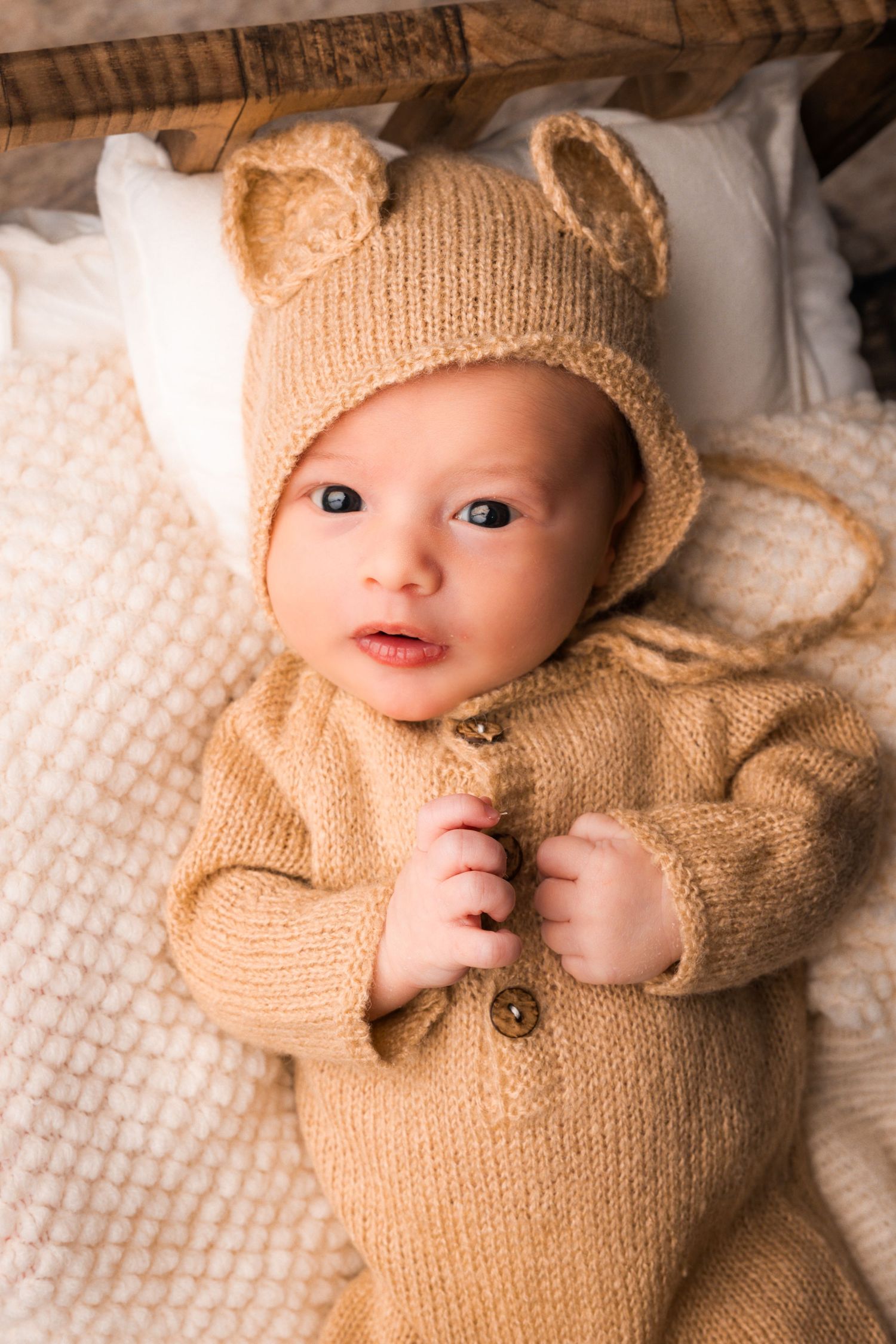 A baby wearing a cozy beige knit sweater lies on white textured bedding.