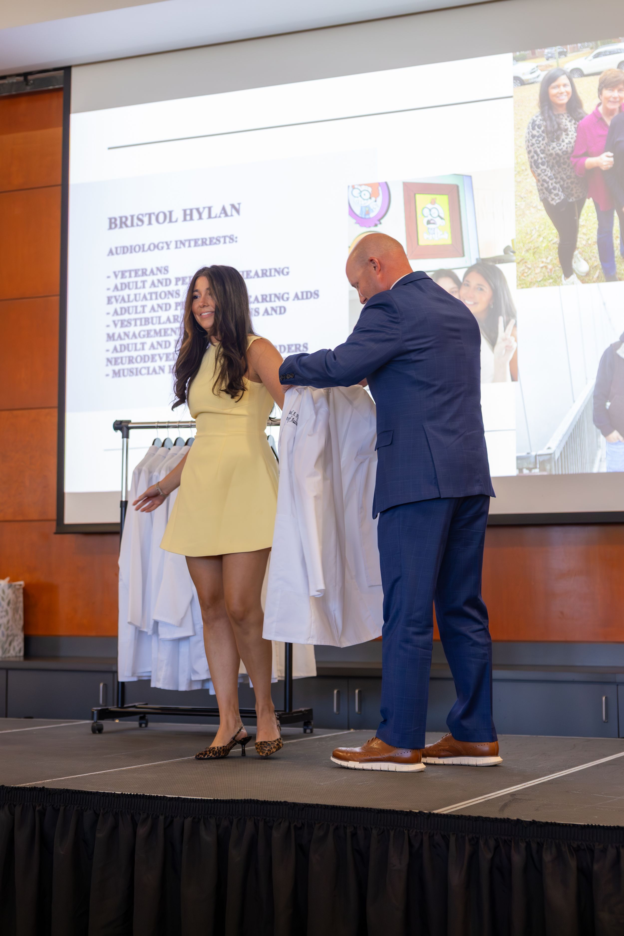 Student receives a white coat during professional ceremony on elevated stage with projection screens in background.