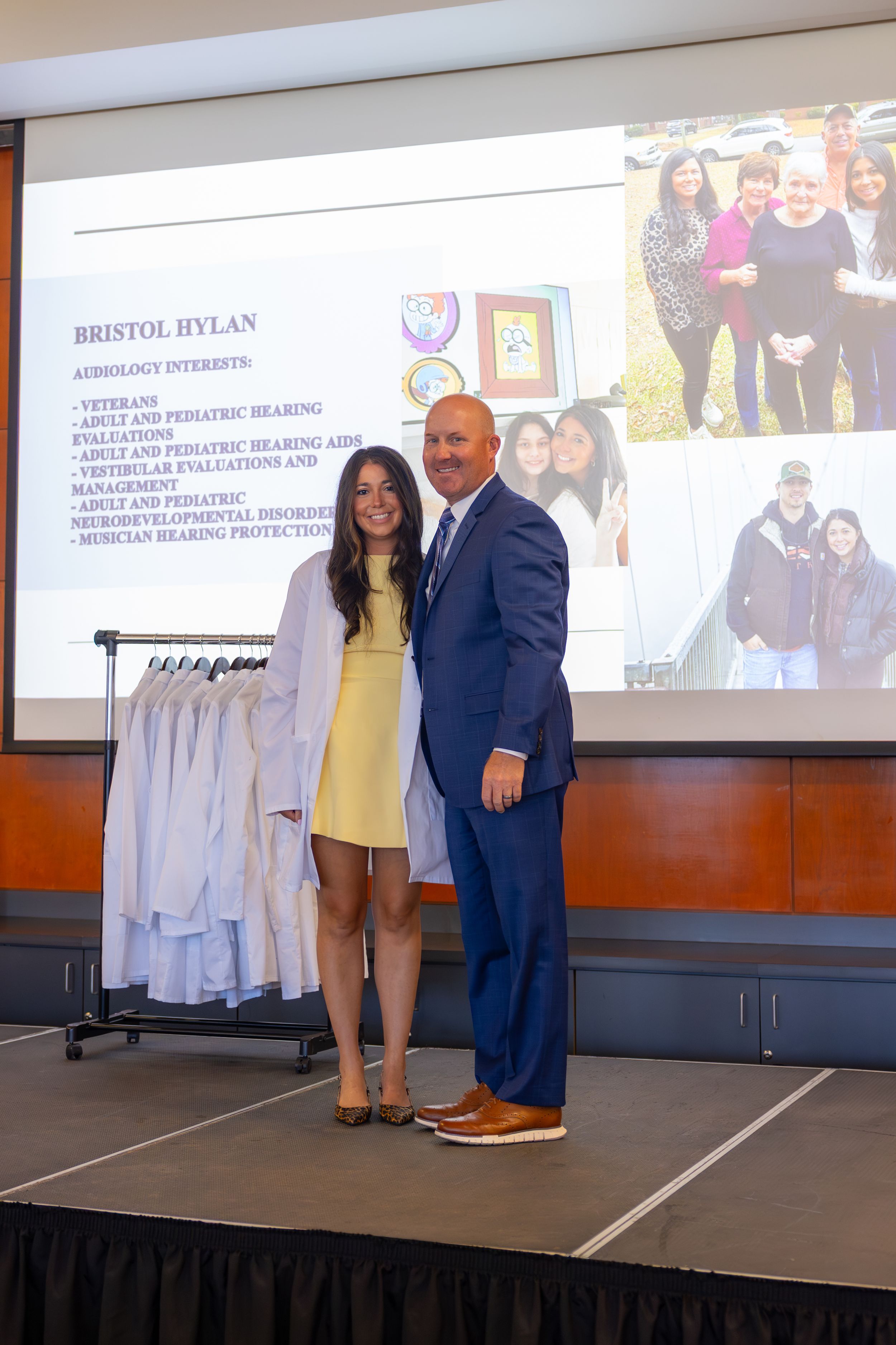 Two people stand on a stage in formal attire with a coat rack and presentation slides visible in the background.