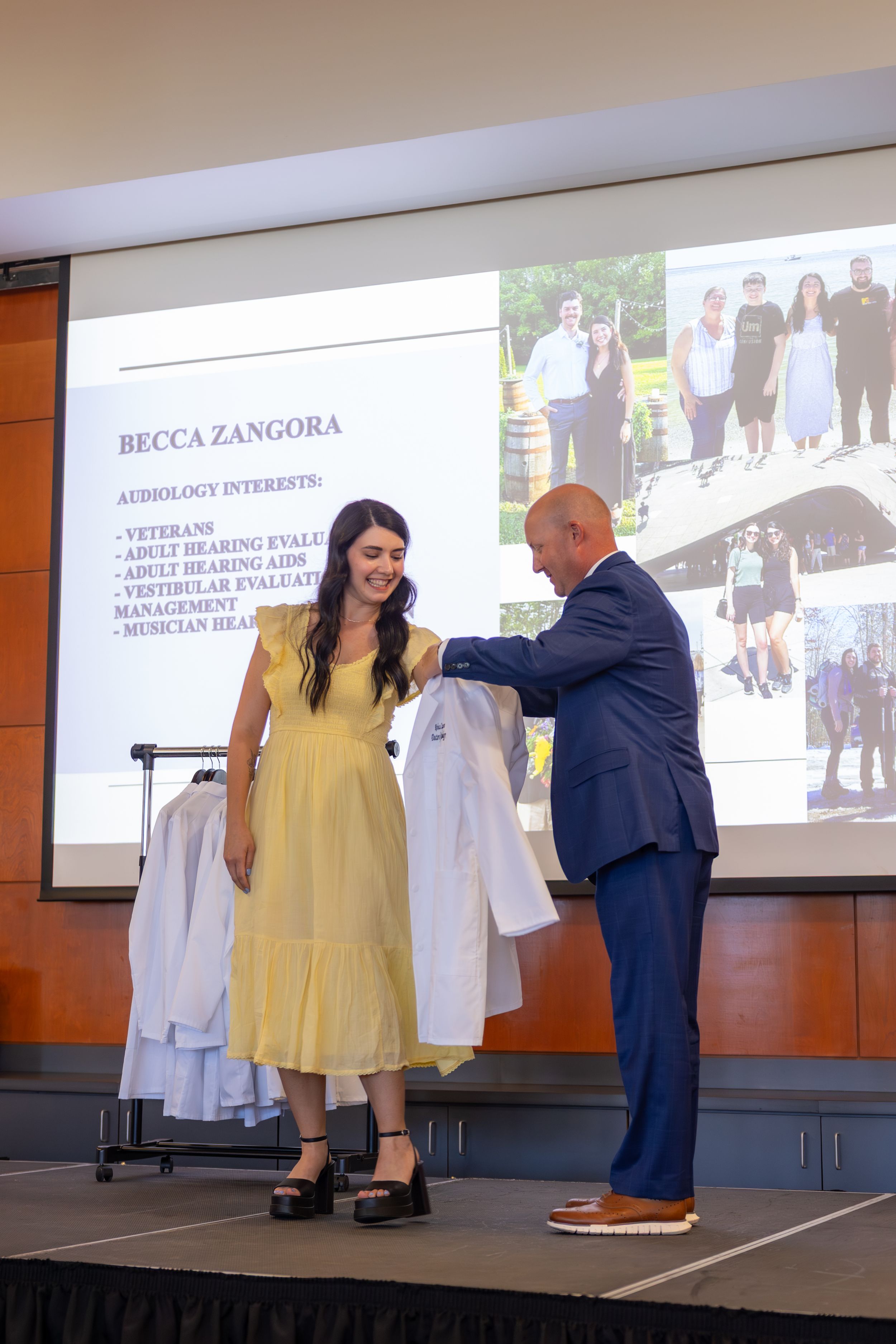 Student in yellow dress receives white medical coat during ceremony on stage with projection screen in background.