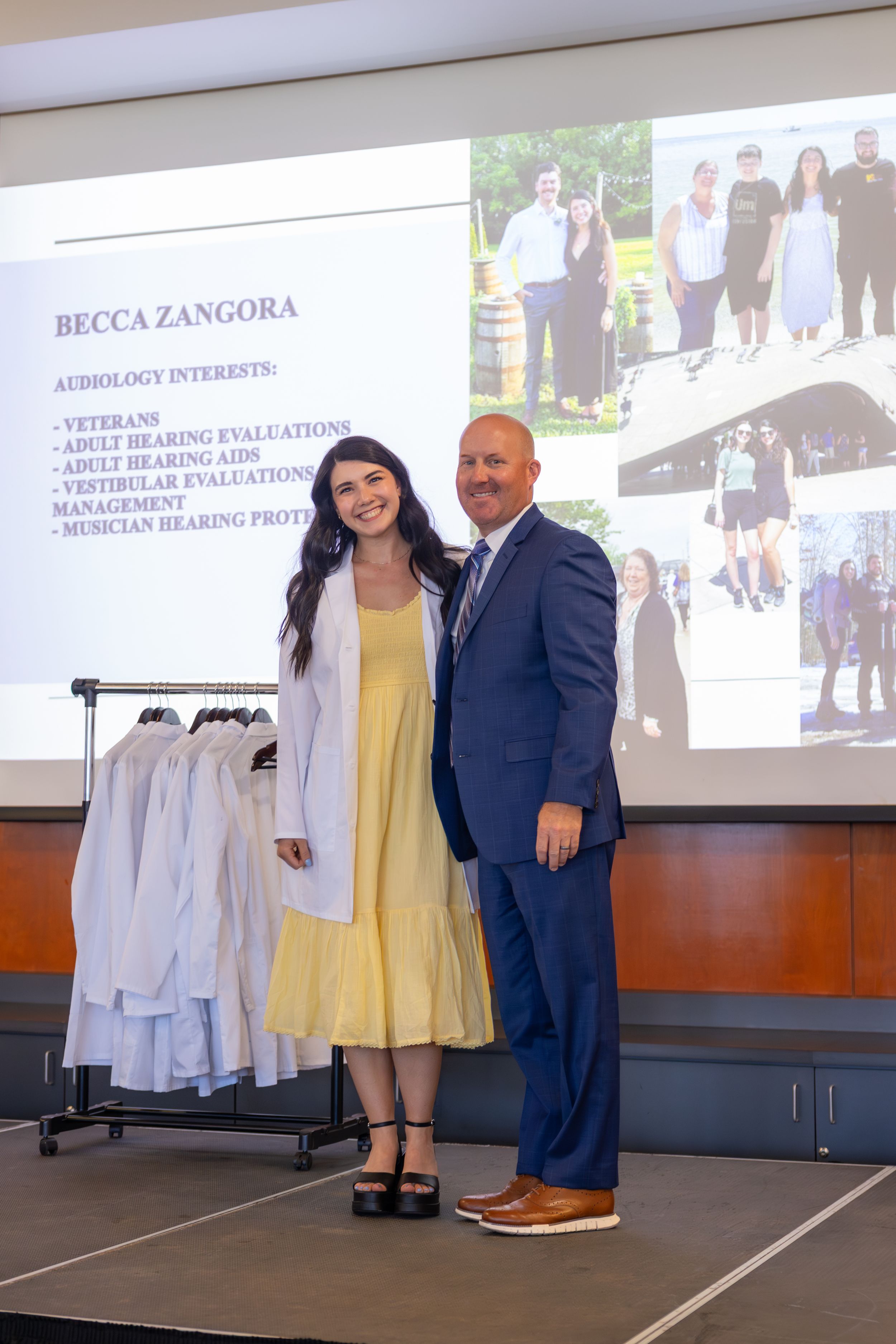 Two professionals pose together in front of a presentation slide showing multiple photos and a clothing rack.