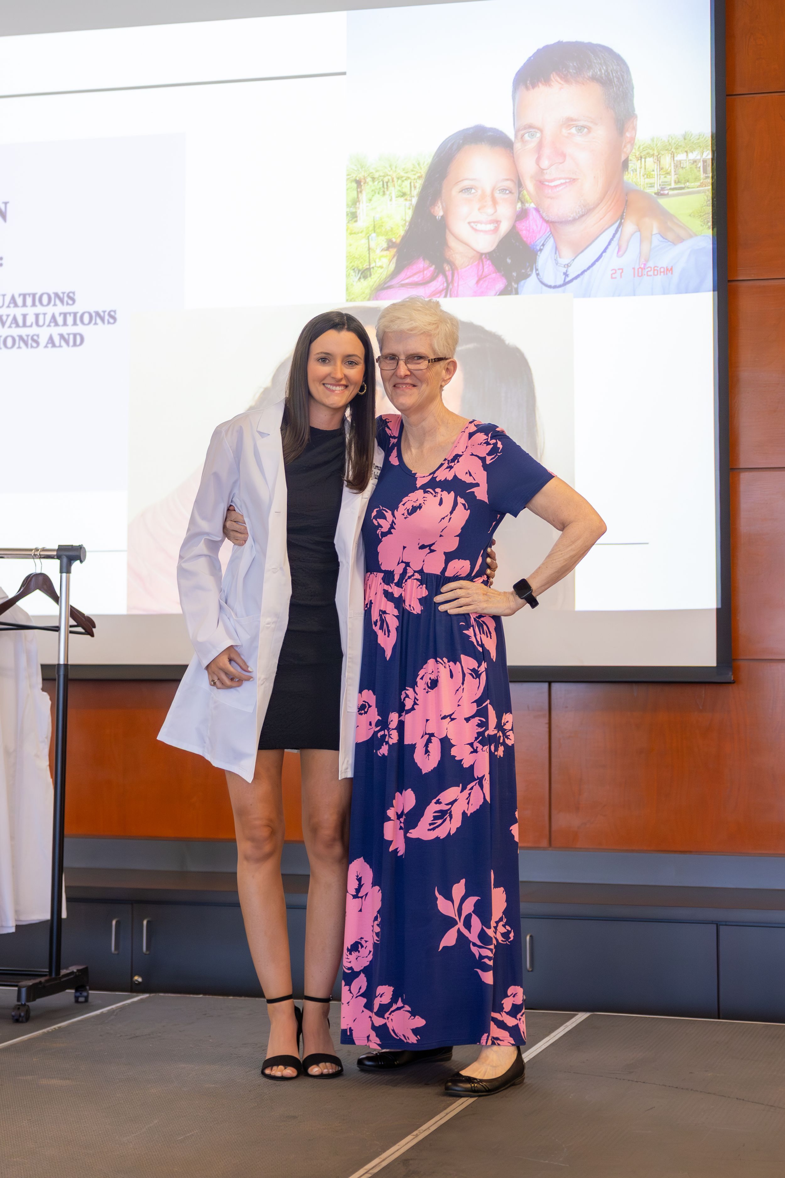 Two women pose together on stage, one in a white coat and black dress, the other in a floral navy maxi dress.