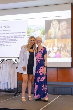 Two people pose together at a presentation event with a projection screen and white clothing rack visible in background.