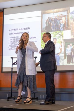Two people standing on stage at a professional presentation with text and photos displayed on screen behind them.