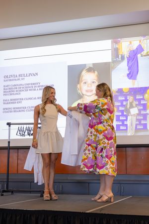 Two people on stage receiving white medical coats during a ceremony with a presentation screen in the background.