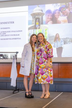 Two individuals pose on stage during a ceremony with photos and presentation slides visible in the background.