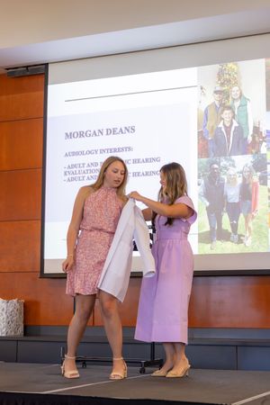 Two people on stage during a white coat ceremony with presentation slides in the background.