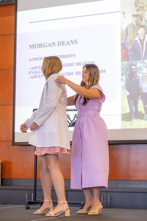 Two medical professionals in white coats participate in a ceremony with projected slides visible.