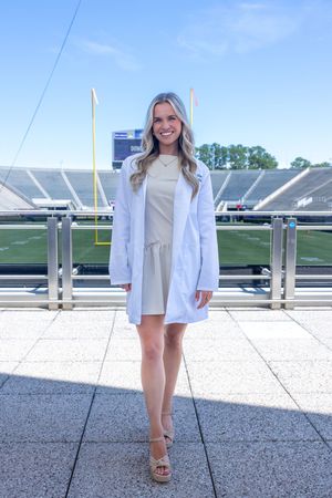 Medical professional in white coat poses at stadium with goal posts visible in background.