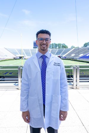 Medical professional in a white coat and blue polka dot tie standing outside a stadium on a sunny day.