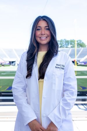 Medical student in white coat stands smiling against football stadium backdrop.