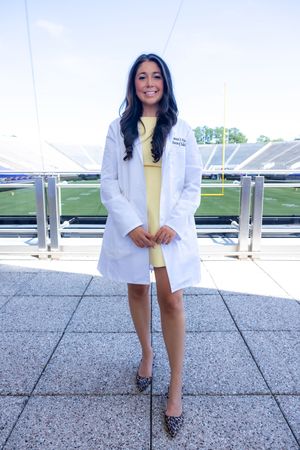 A person in a white medical coat and yellow dress poses at a sports stadium venue.