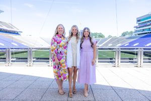 Three people wearing summer dresses pose together at a stadium with bleachers visible in the background.