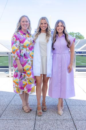 Three people in colorful dresses pose together on a stadium concourse during a sunny day.