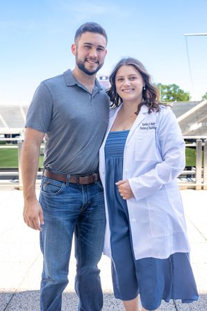 Medical student in white coat poses with supporters for graduation photos outside a stadium.