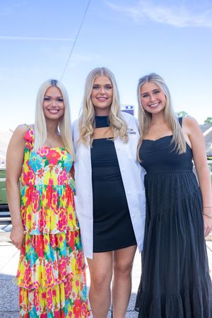 Three women celebrating at a medical school ceremony, wearing colorful dresses and white coat.