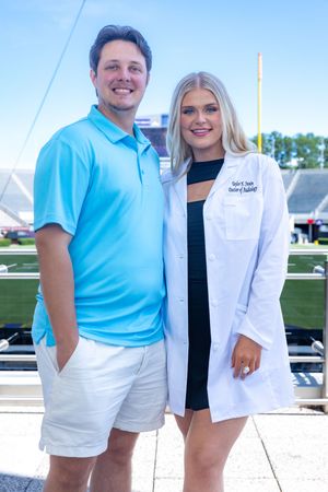 Couple poses together at medical school ceremony with football field in background.