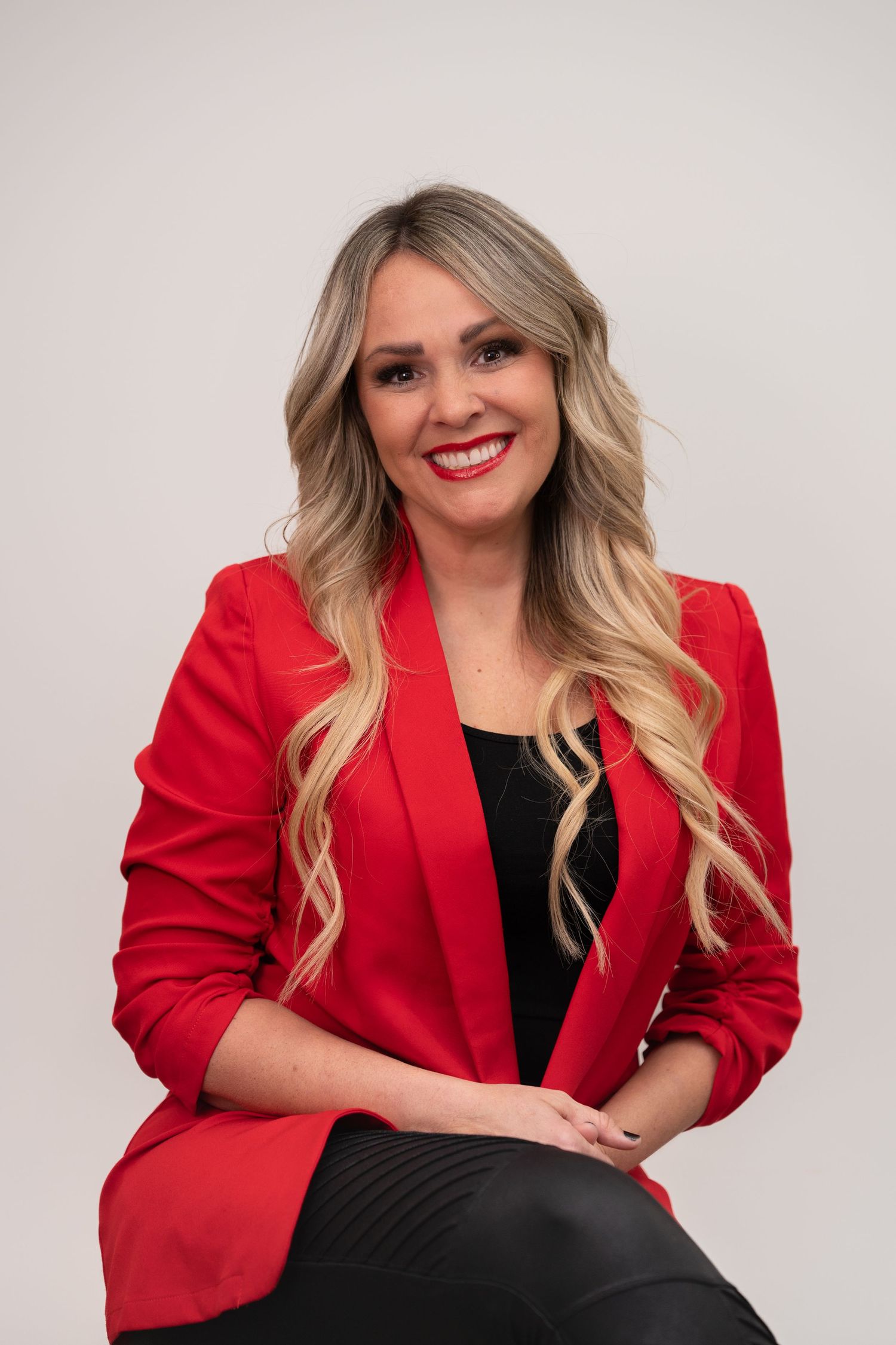 Professional headshot of someone in a bright red blazer and black top with long wavy blonde hair against light background.