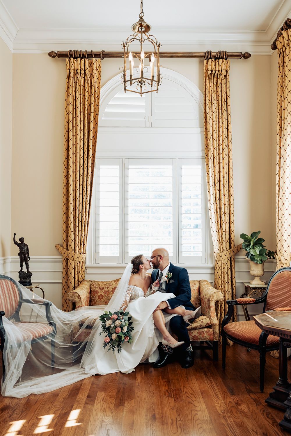 Romantic wedding photo with elegant gold curtains, hardwood floors, arched window and ornate lantern chandelier hanging from white ceiling.