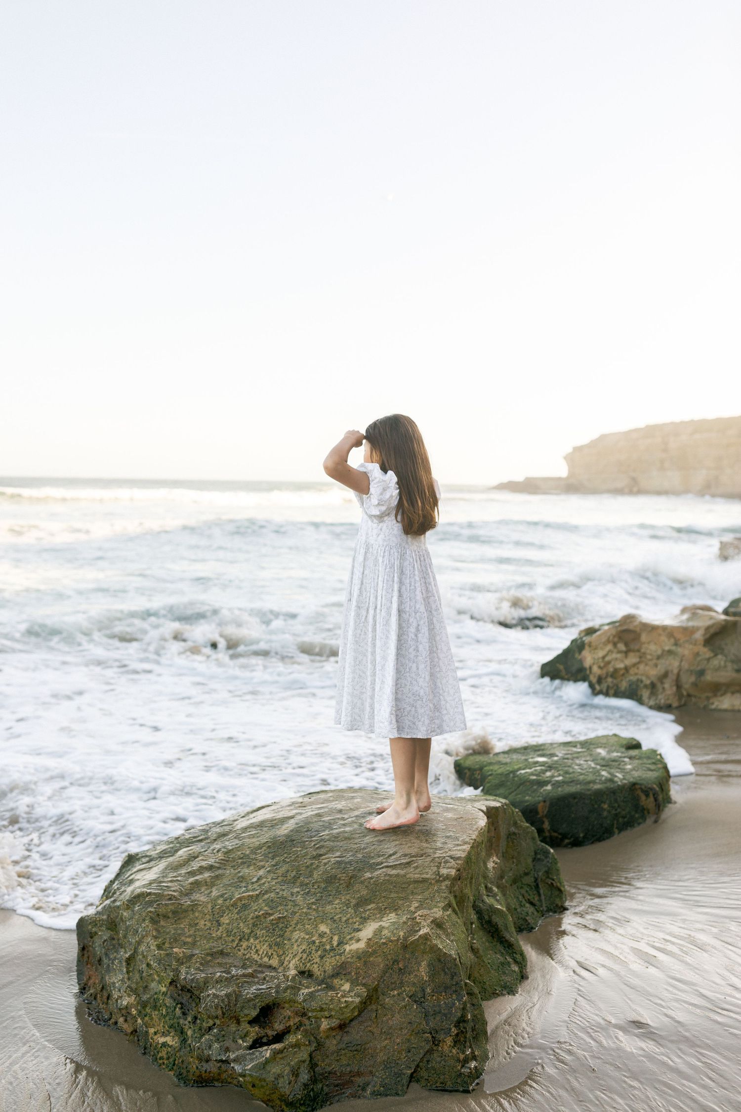 A person in a white sundress stands on a coastal rock formation looking out at ocean waves during sunset.