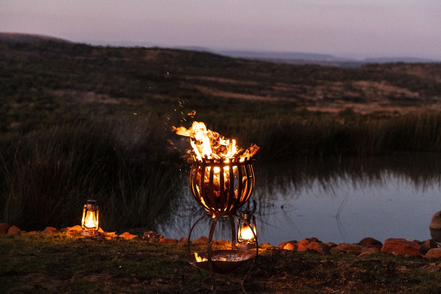 Experience a fire burning at dusk beside tranquil waters in African bush at a Luxury Safari Lodge.