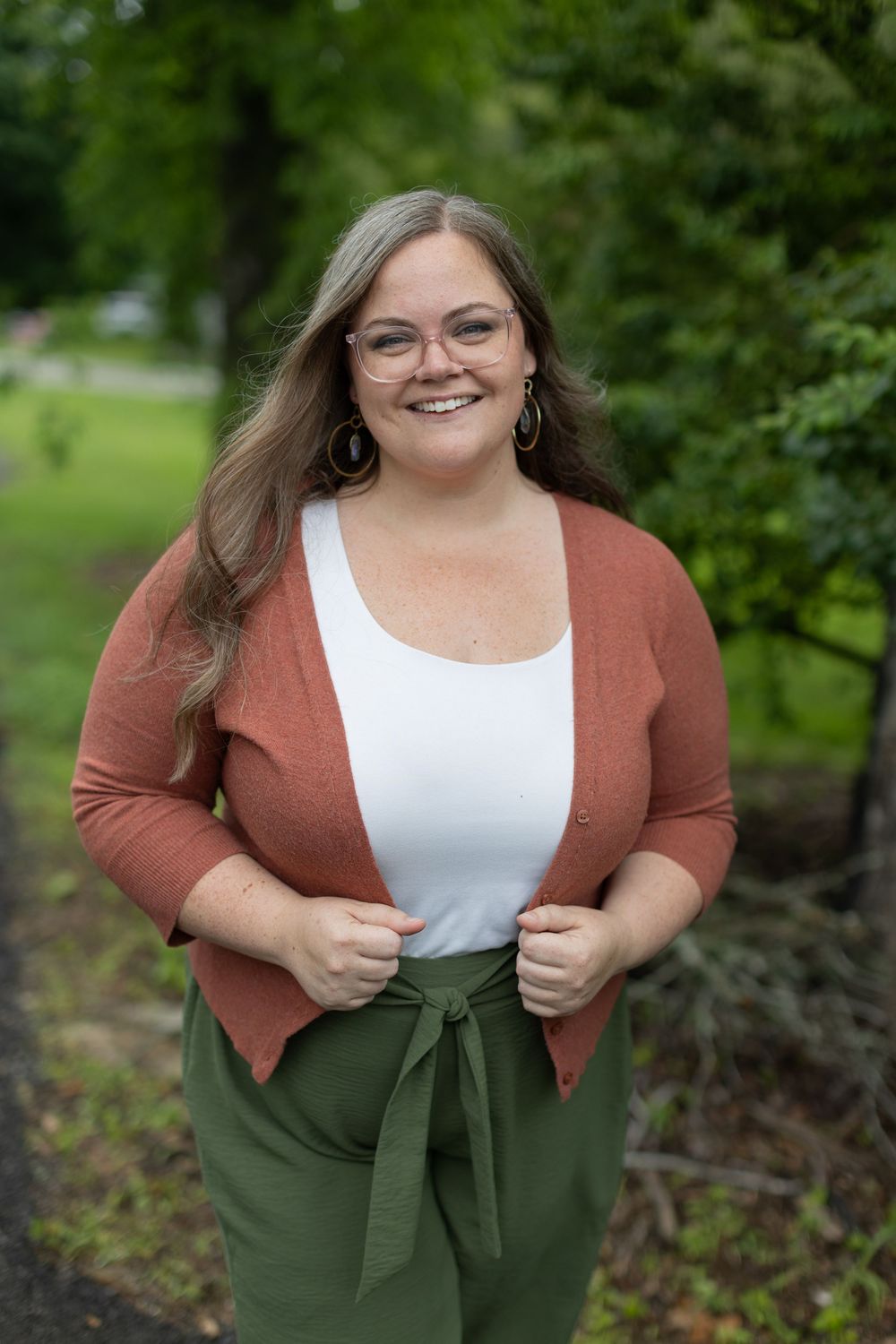 A person wearing a rust-colored cardigan, white top, and olive green skirt poses outdoors in a natural setting.