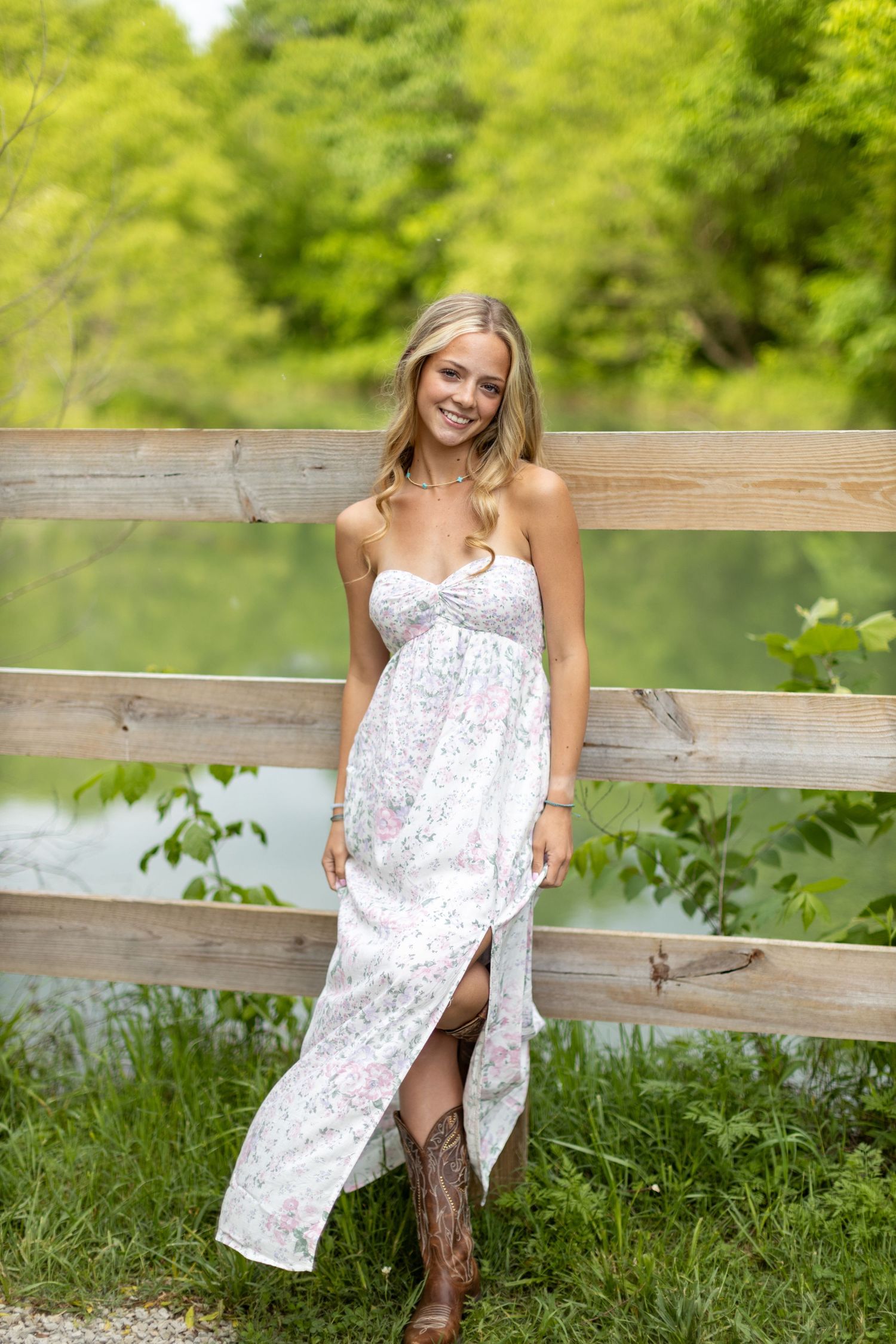 Summer portrait of someone wearing a white floral maxi dress and cowboy boots standing by a rustic wooden fence near water.