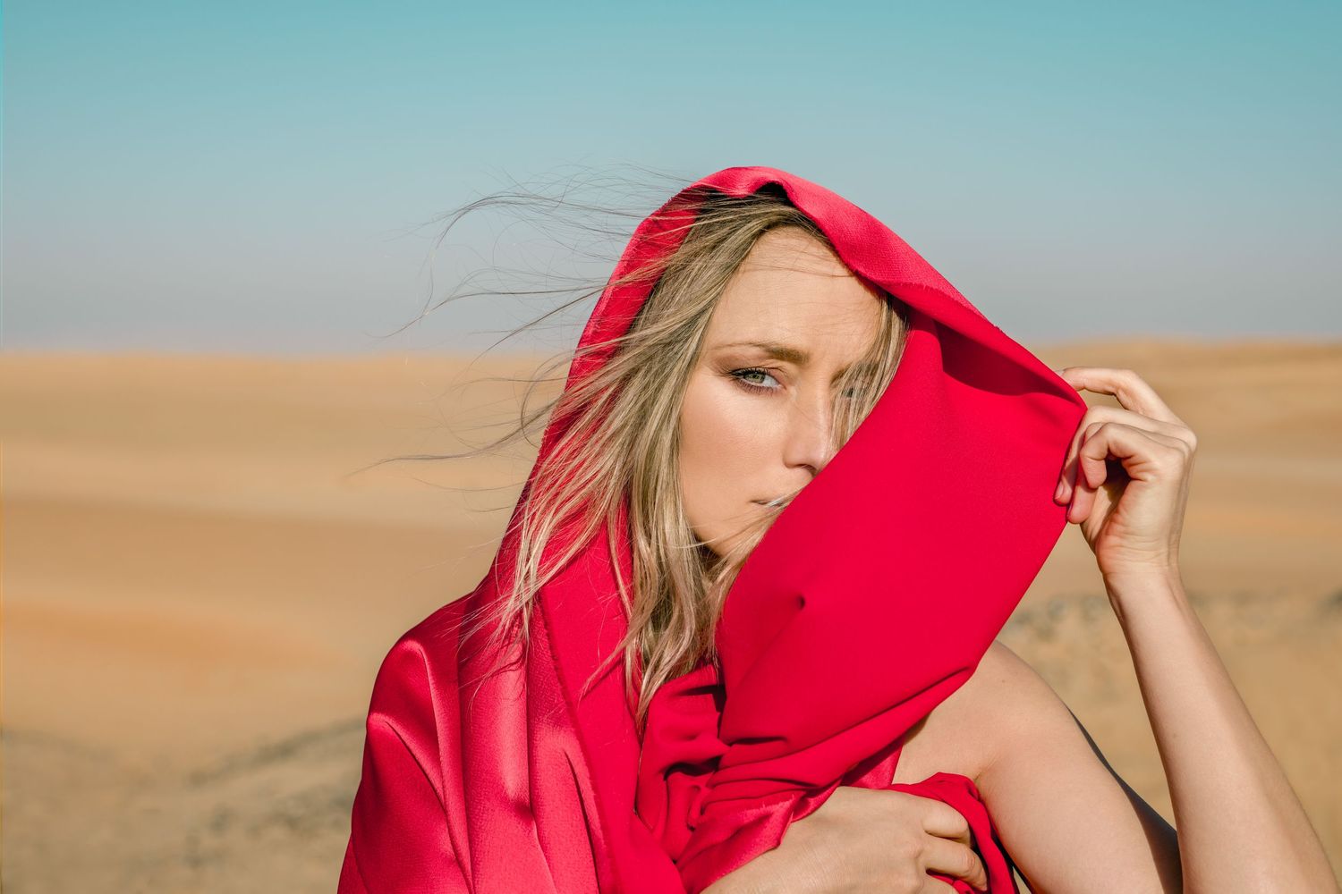 A model in a flowing red dress poses dramatically against sandy desert dunes at sunset. Work from The Moment of endless wind, exhibition.