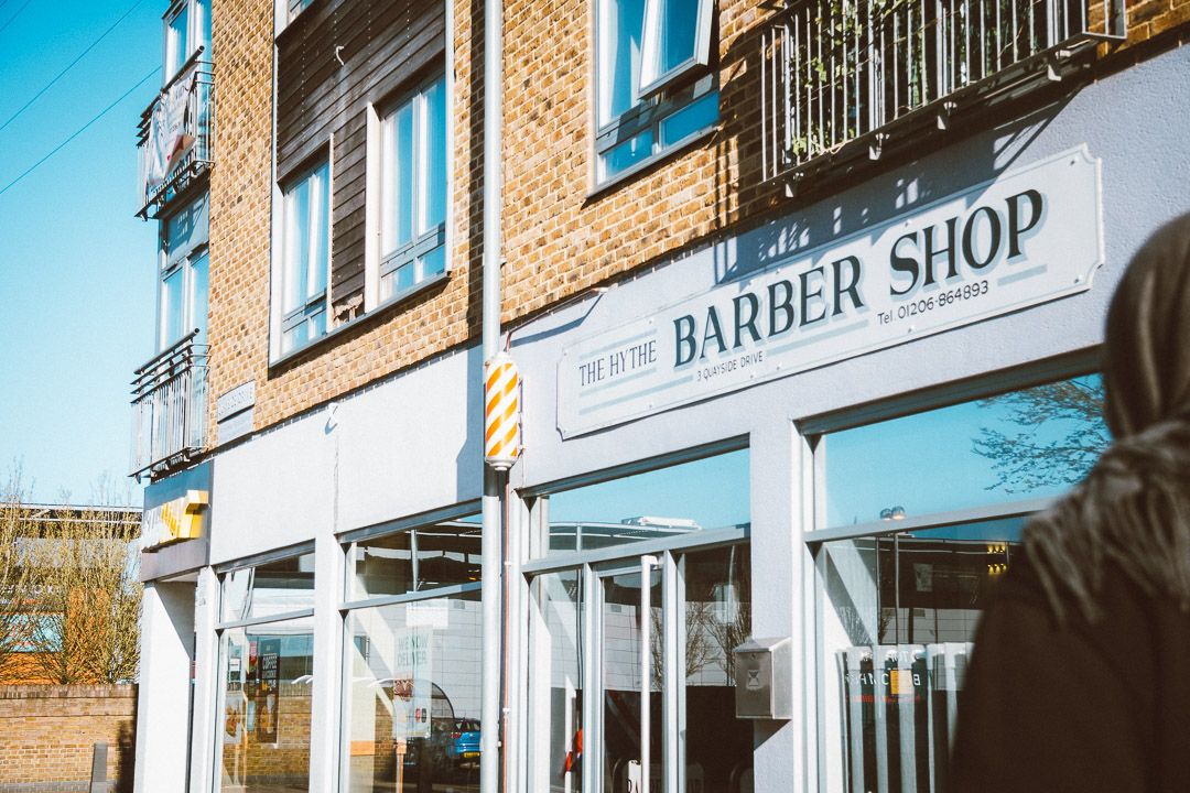 A storefront exterior view of a barber shop with brick walls and large glass windows on a sunny day.