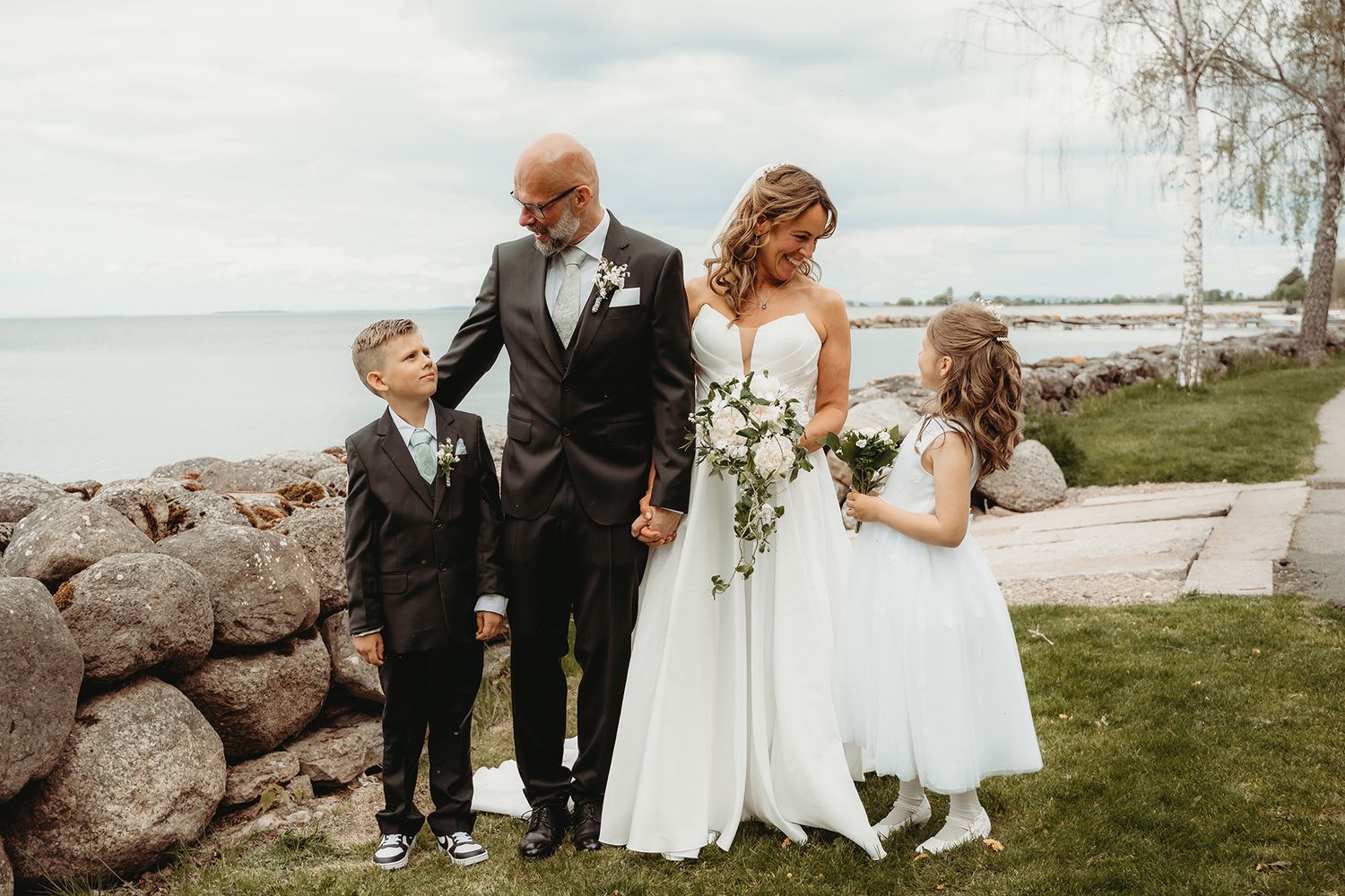 Familjeporträtt på bröllop, brudparet står med sina två smp barn intill Vätterns strand längs en lång stenmur i Vadstena.