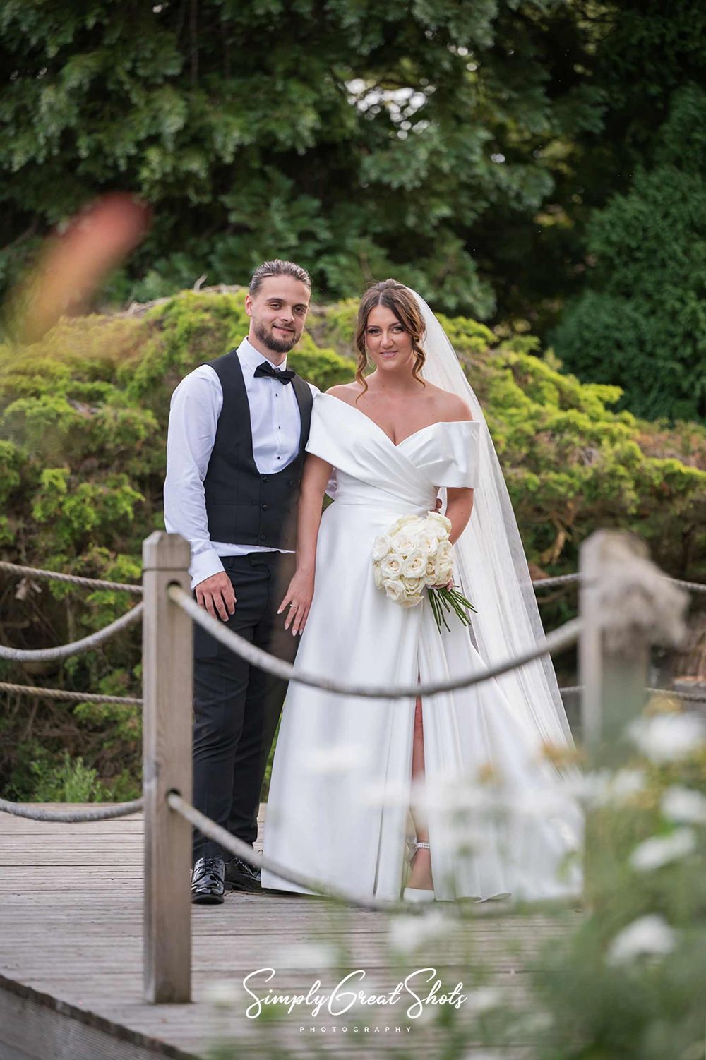 An elegant wedding couple in formal attire pose for wedding photos on a wooden bridge in a garden setting with white floral bouquet.