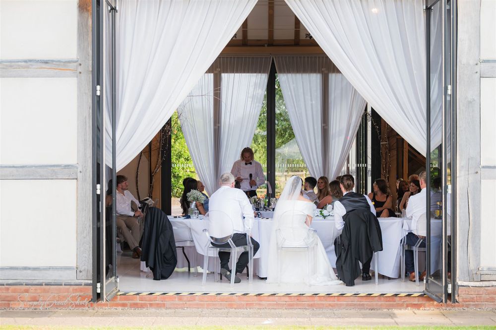 The best man speech with wedding guests seated at tables inside an elegant venue with white curtains & open barn doors framing the entrance