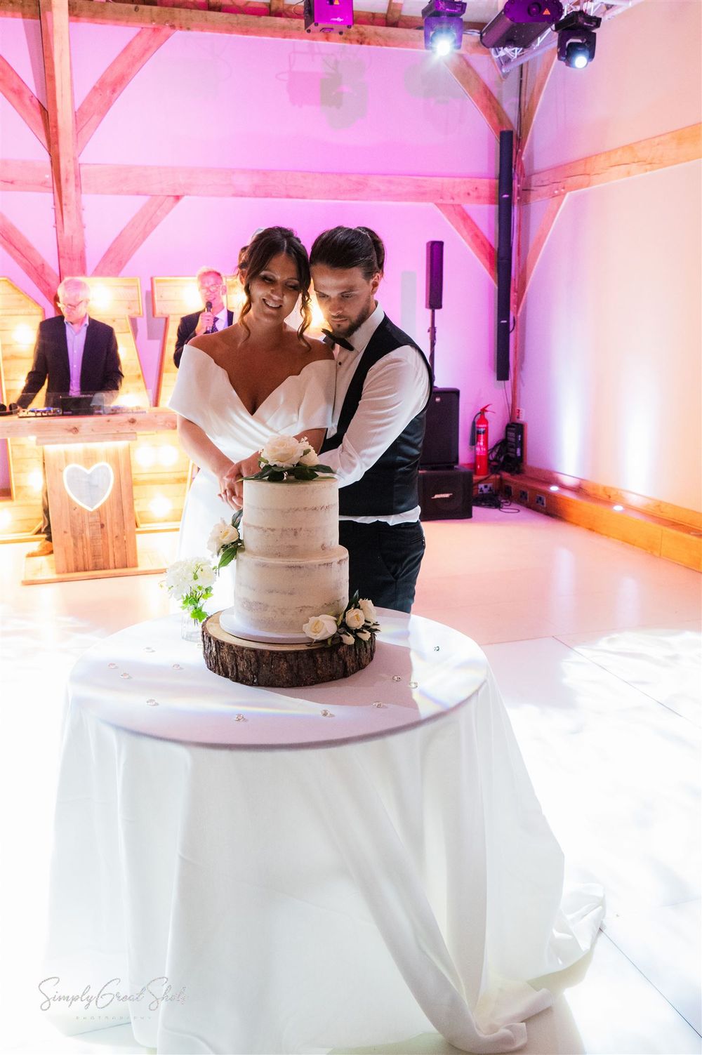 A bride and groom cut their elegant white wedding cake together at a reception with pink mood lighting at Redhouse Barn wedding venue