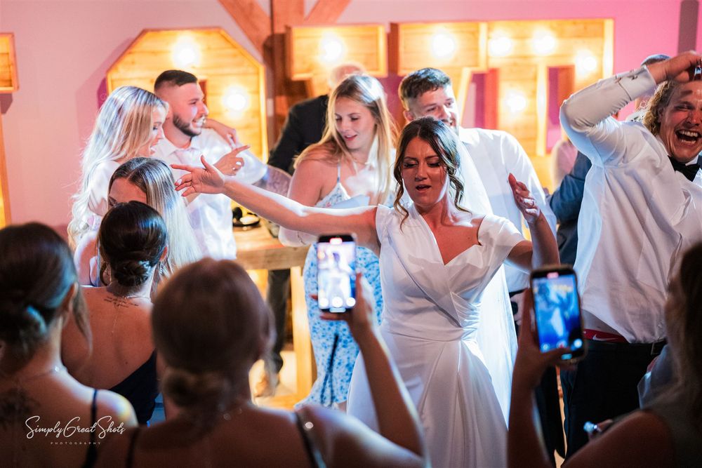 The bride and her guests celebrate and dance together at a wedding reception with warm lighting and glowing orange decor in the background.