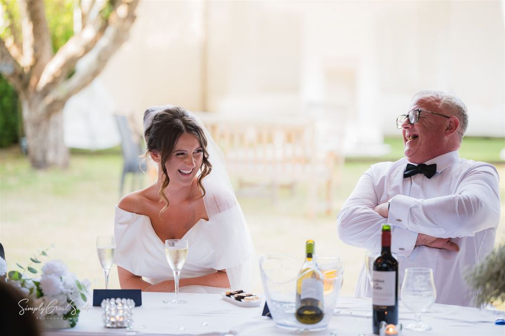 The bride & father-of-the-bride share a laugh while seated at a reception table with doors open to the outside gardens with bottles of wine.