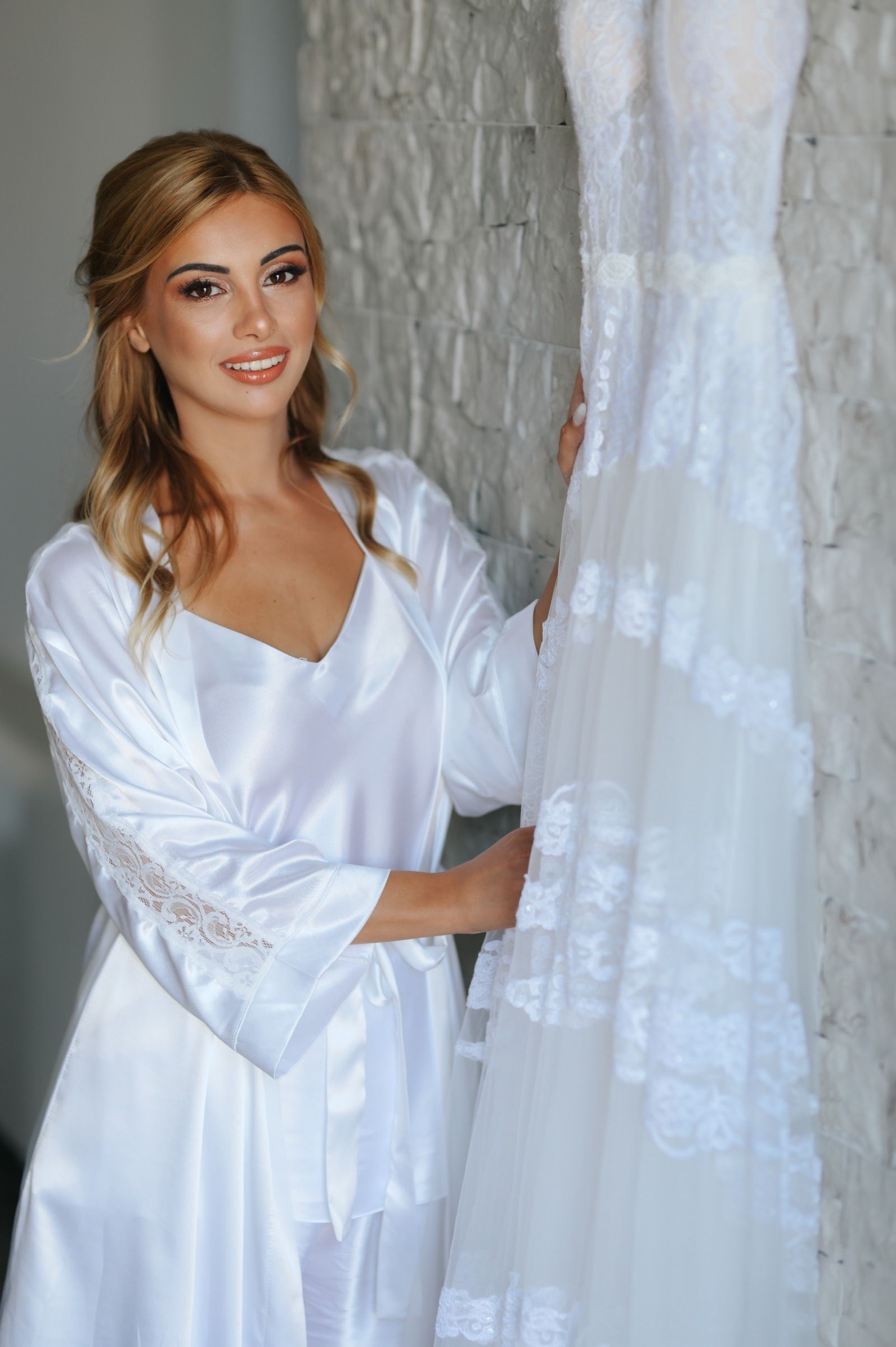 Woman in white silk robe smiles while standing next to hanging white lace wedding dress in bridal preparation room.