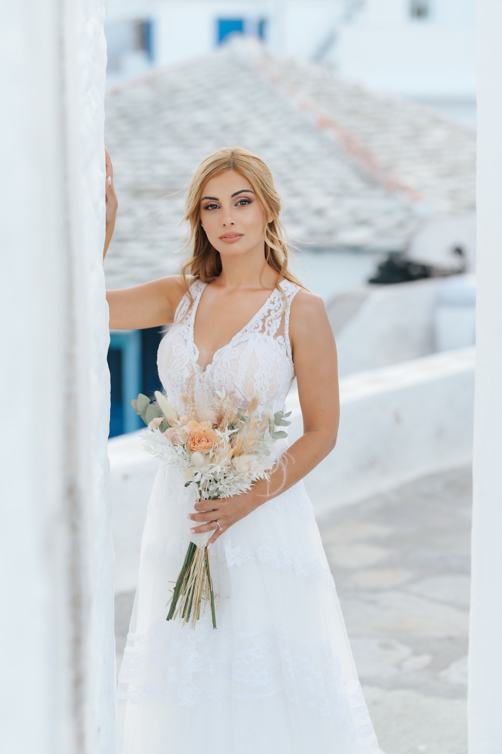 Elegant bridal portrait featuring a white sleeveless wedding gown and delicate peach floral bouquet against a white Mediterranean backdrop.