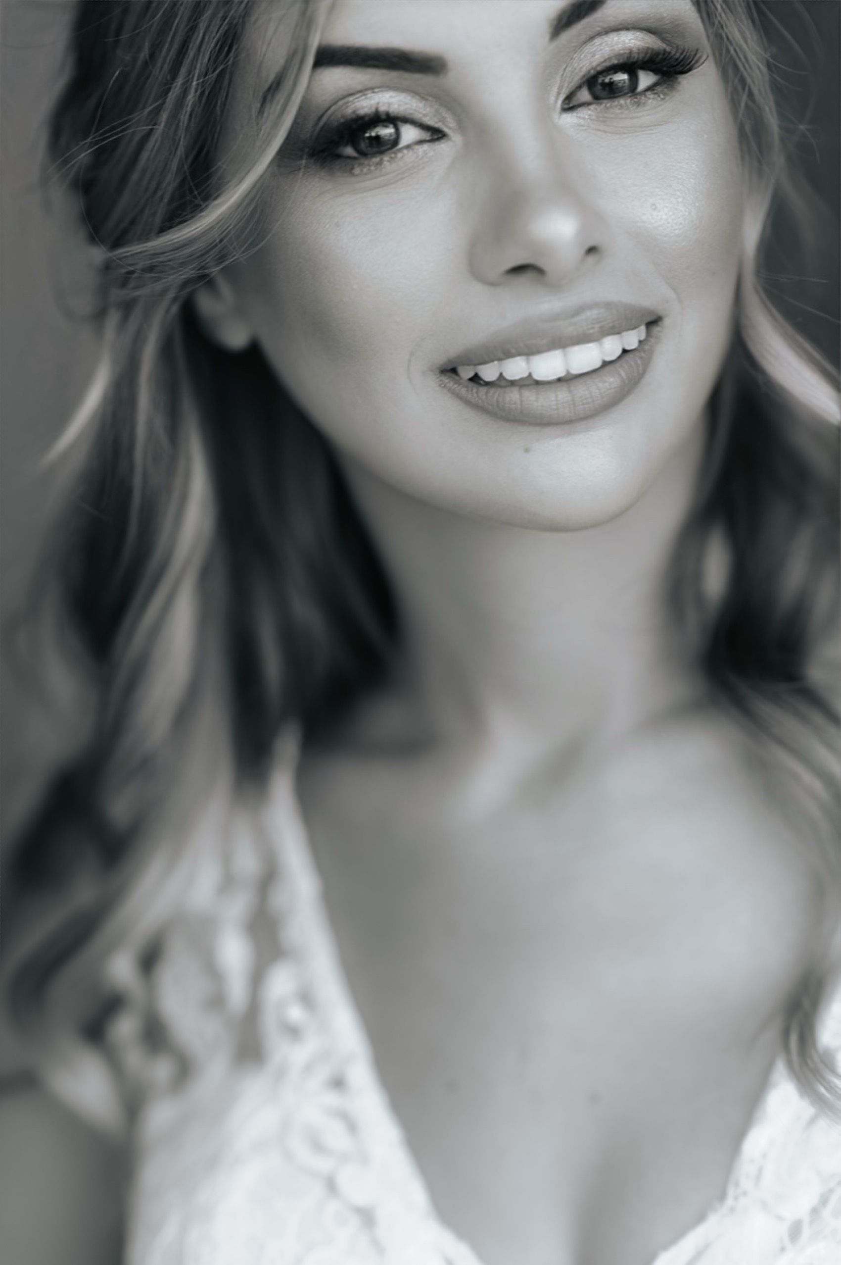 Black and white portrait shows a smiling model with long hair wearing a lace top in soft lighting.