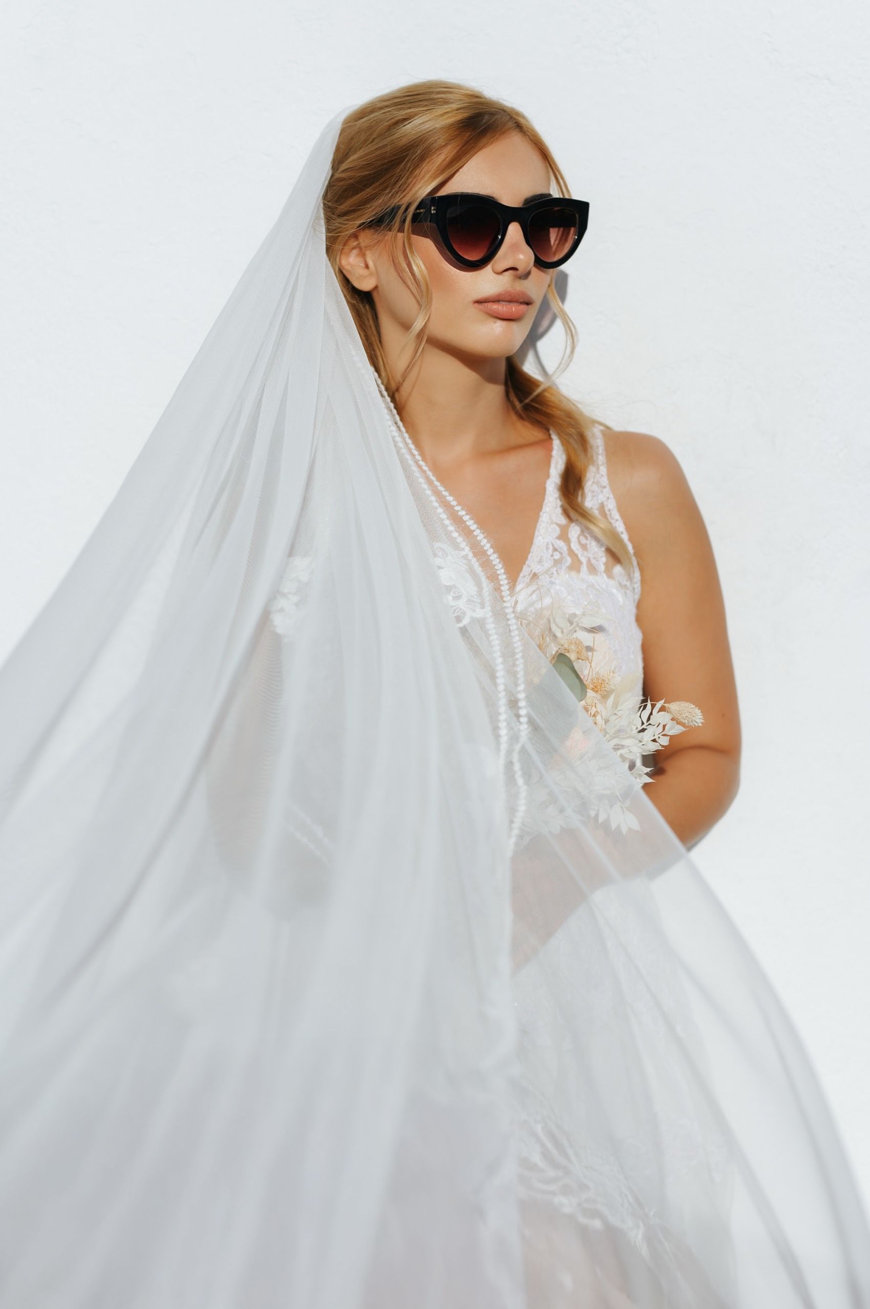 A bride in a white lace wedding gown and sunglasses poses with flowing veil against a light background.