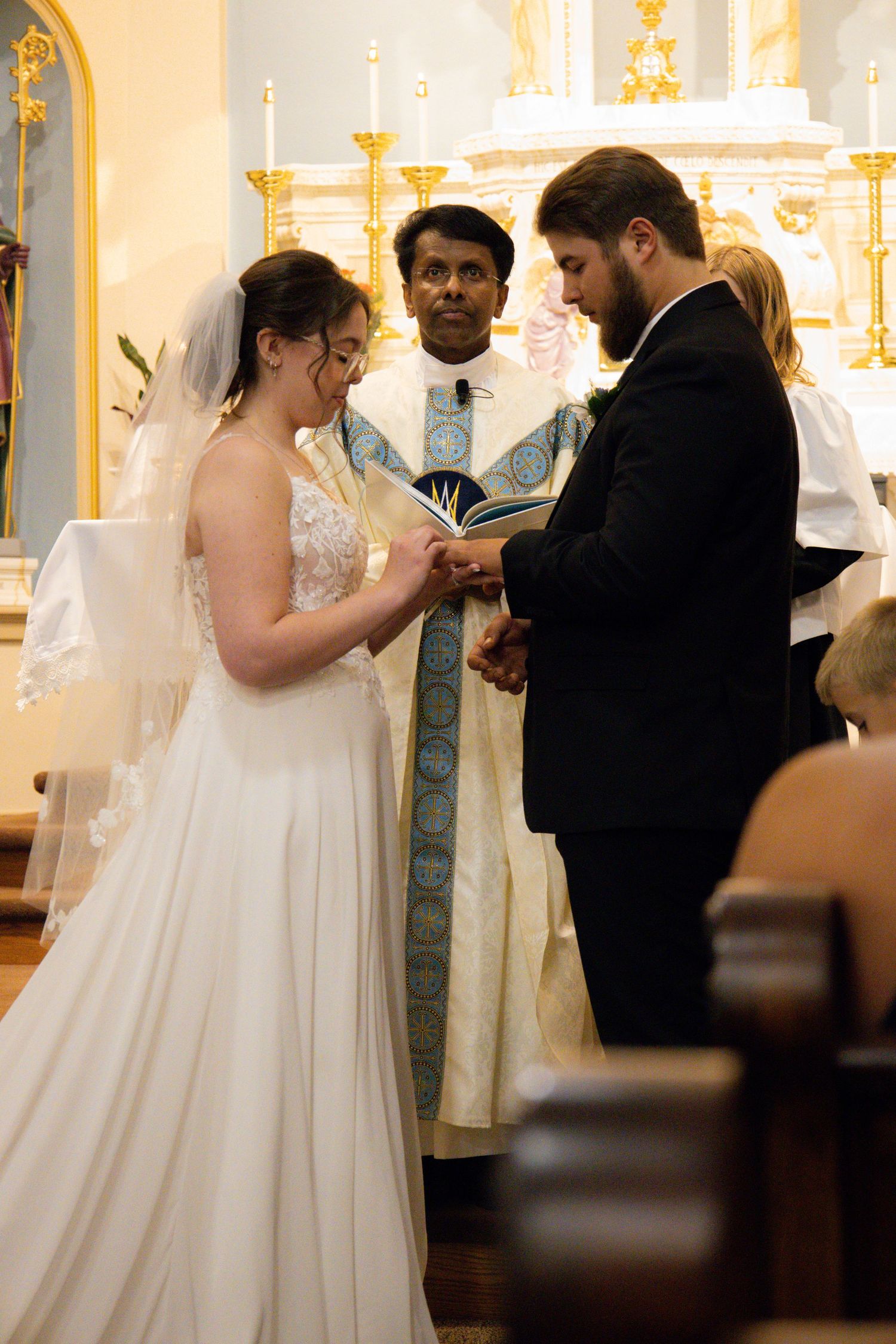 A wedding ceremony takes place at the altar in an ornate church with a priest in light blue vestments presiding.