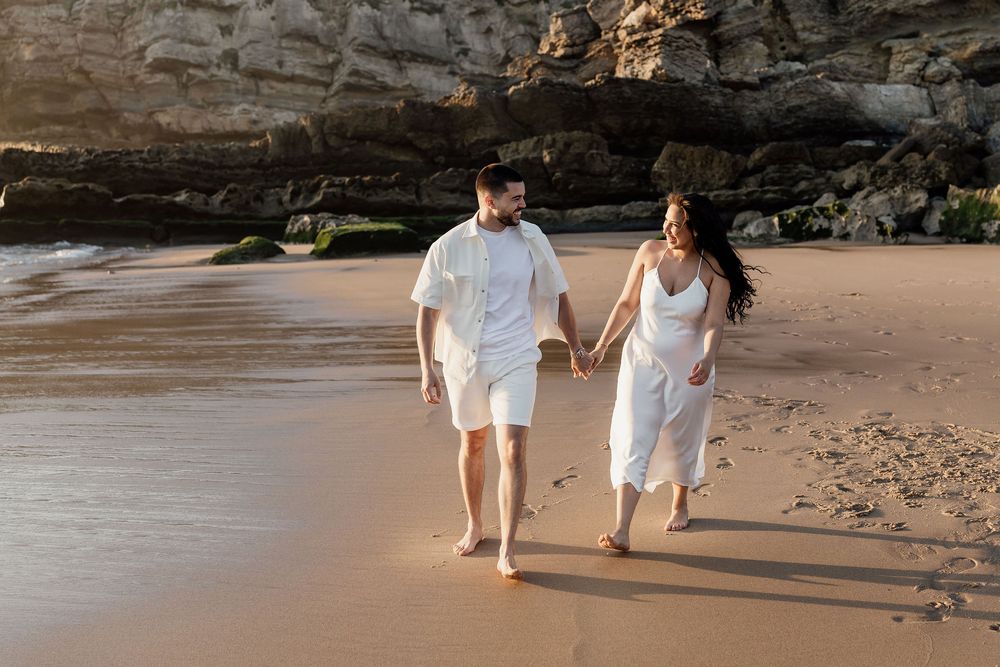 Couple in white outfits walking barefoot along a scenic beach at sunset with cliffs in the background.