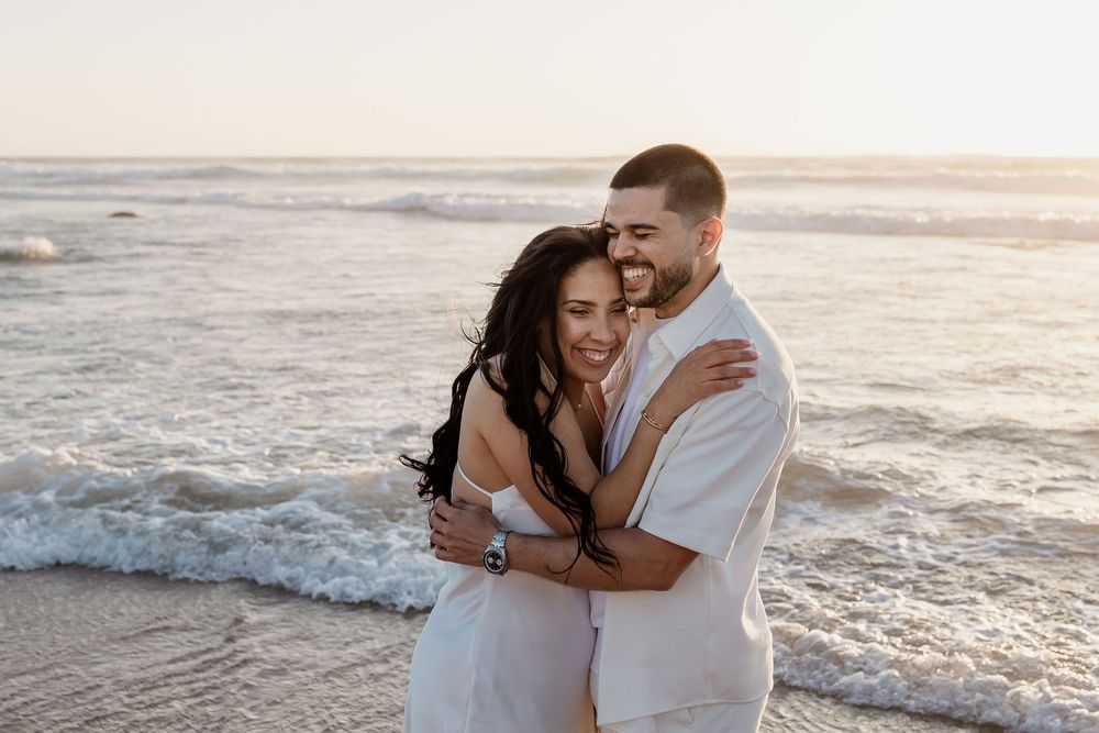 A happy couple embraces on a sandy beach at sunset while waves crash behind them.