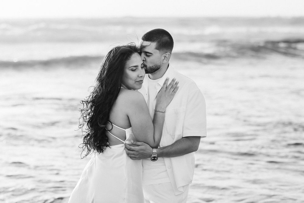 A romantic black and white photo of a couple embracing on the beach with ocean waves in the background.