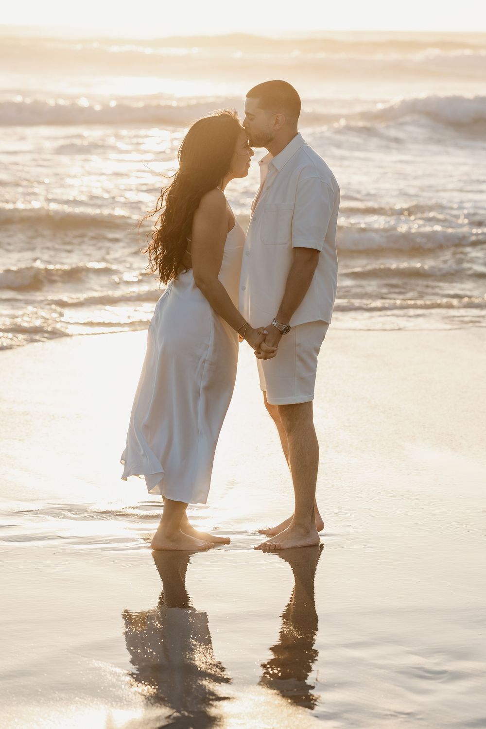 A loving couple stands barefoot in shallow beach water at sunset, creating a romantic coastal silhouette.