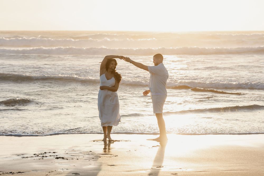 Two figures dancing together on a sunlit beach at golden hour as ocean waves roll in along the shoreline.